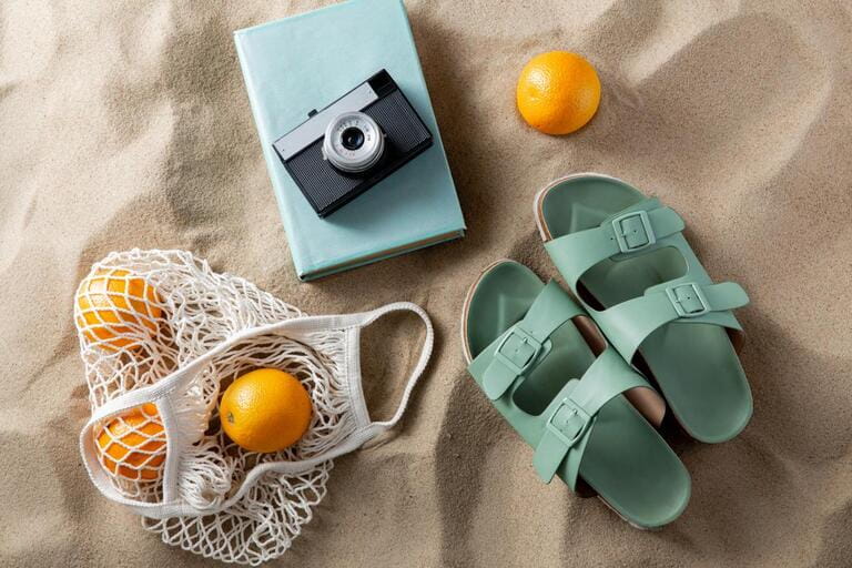 leisure and summer holidays concept - slippers, string bag of oranges, film camera and book on beach sand