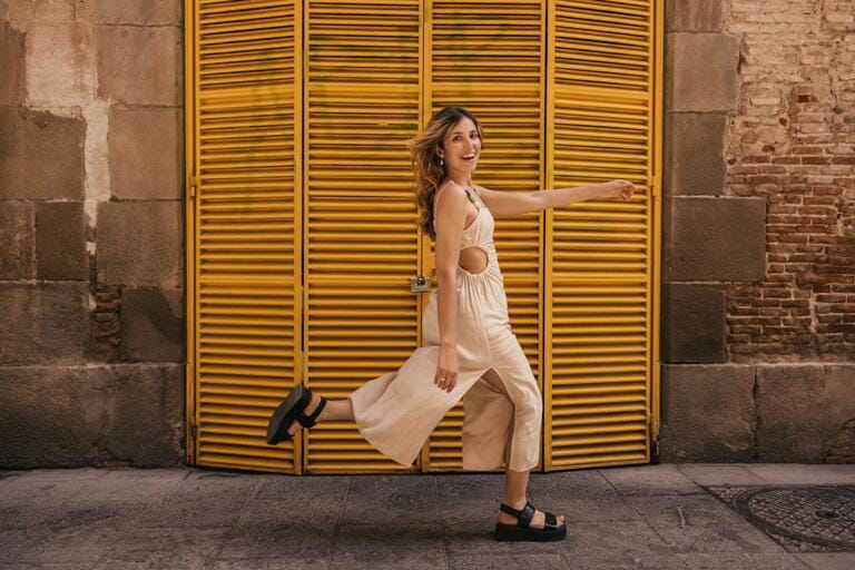 Funny young caucasian woman looks at camera, runs along street in warm weather on weekend. Blonde wears beige sundress and black sandals. Summer vacation concept