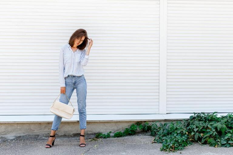Fashionable young woman wearing blue striped shirt, blue cropped denim jeans, black high heel sandals and beige handbag posing outdoors against white street wall. Fashionable casual outfit. Street fashion.