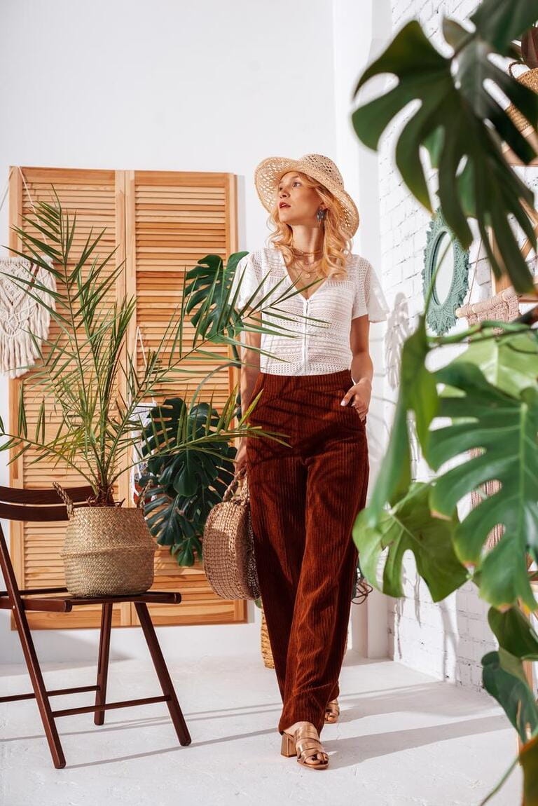 Full-lengh indoor portrait of fashionable woman wearing summer white crochet top, brown corduroy trousers, straw hat, sandals, posing at home, in stylish boho interior with green tropical plants