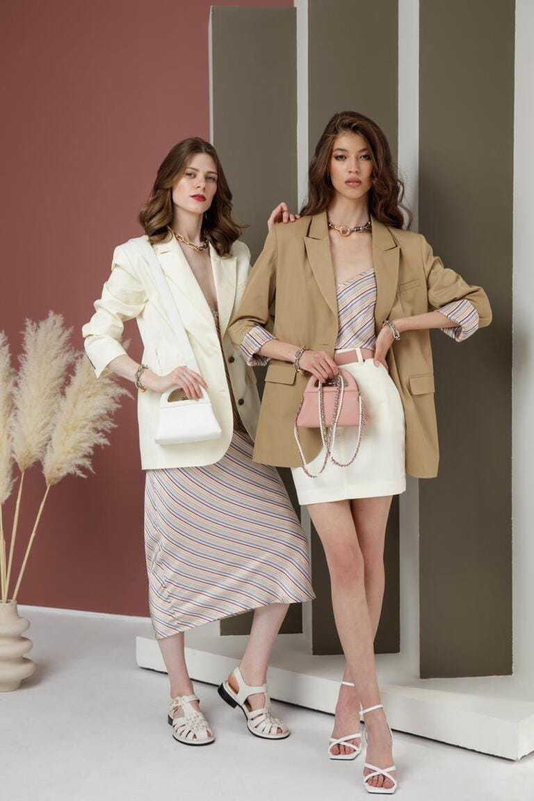 Two fashion models in white cream and sand brown looks. Jacket, blazer, skirt, long dress, handbag. Beautiful young women. Rosy brown and Striped Corrugated Wall, dry branches. Asian, european girls