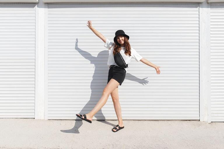 Woman, wearing white t-shirt, black shorts, fanny pack or waist pack, bucket hat and flat sandals, standing outdoor near white wall. Stylish trendy basic minimalistic casual outfit. Street fashion Woman, wearing white t-shirt, black shorts, fanny pack or waist pack, bucket hat and flat sandals, standing outdoor near white wall. Stylish trendy basic minimalistic casual outfit. Street fashion