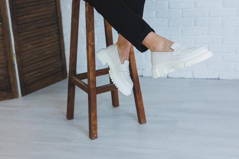 Legs of a young beautiful woman in black trousers and modern stylish white leather loafers. New collection of women's shoes spring-summer 2022