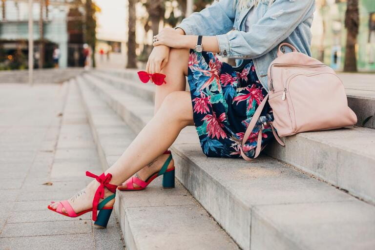 close up details of legs in pink sandals of woman sitting on stairs in city street in stylish printed skirt with leather backpack holding sunglasses, summer style trend