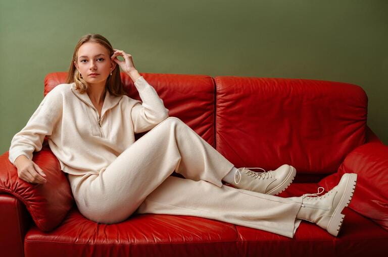 Fashionable confident woman wearing elegant white knit suit, with zip neck sweater, wide leg trousers, leather ankle lace up boots, sitting, posing on leather couch. Fashionable confident woman wearing elegant white knit suit, with zip neck sweater, wide leg trousers, leather ankle lace up boots, sitting, posing on leather couch.