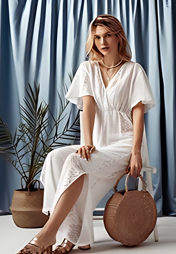 Fashionable blonde woman wearing trendy summer crochet jumpsuit, layered pearl necklace, sandals, holding round straw wicker bag. Full-length indoor portrait